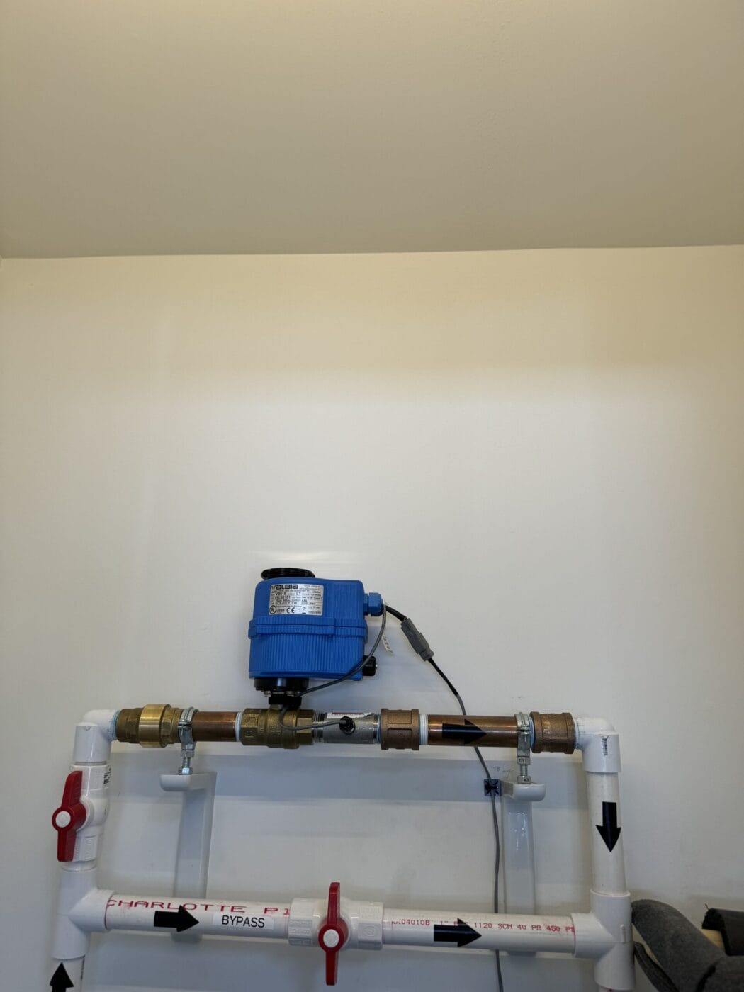 A plumbing setup with a blue control device mounted on a wall. Two white pipes with red handles and black directional arrows are below the device.