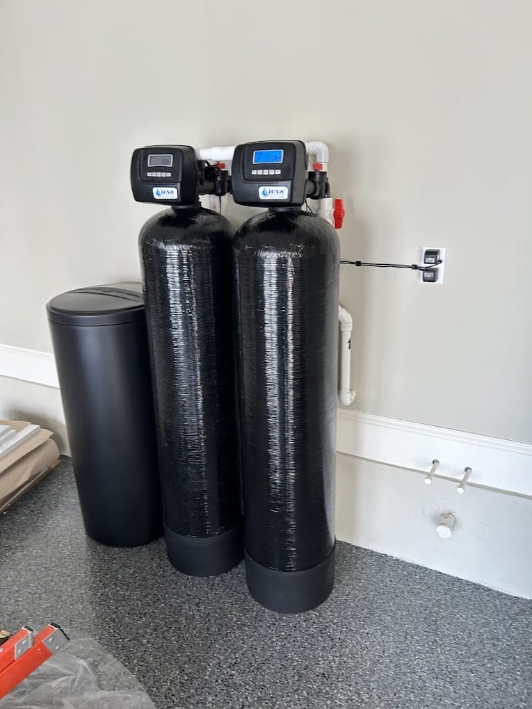 Two tall black water softener tanks with digital controls are positioned next to a black cylindrical container on a speckled floor.