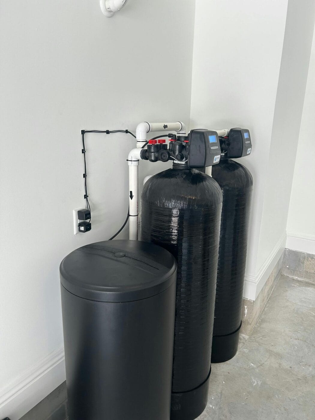 Two large black water softener tanks with digital display controls are positioned against a wall, connected by white pipes for whole house water purification. A round black brine tank sits next to them.